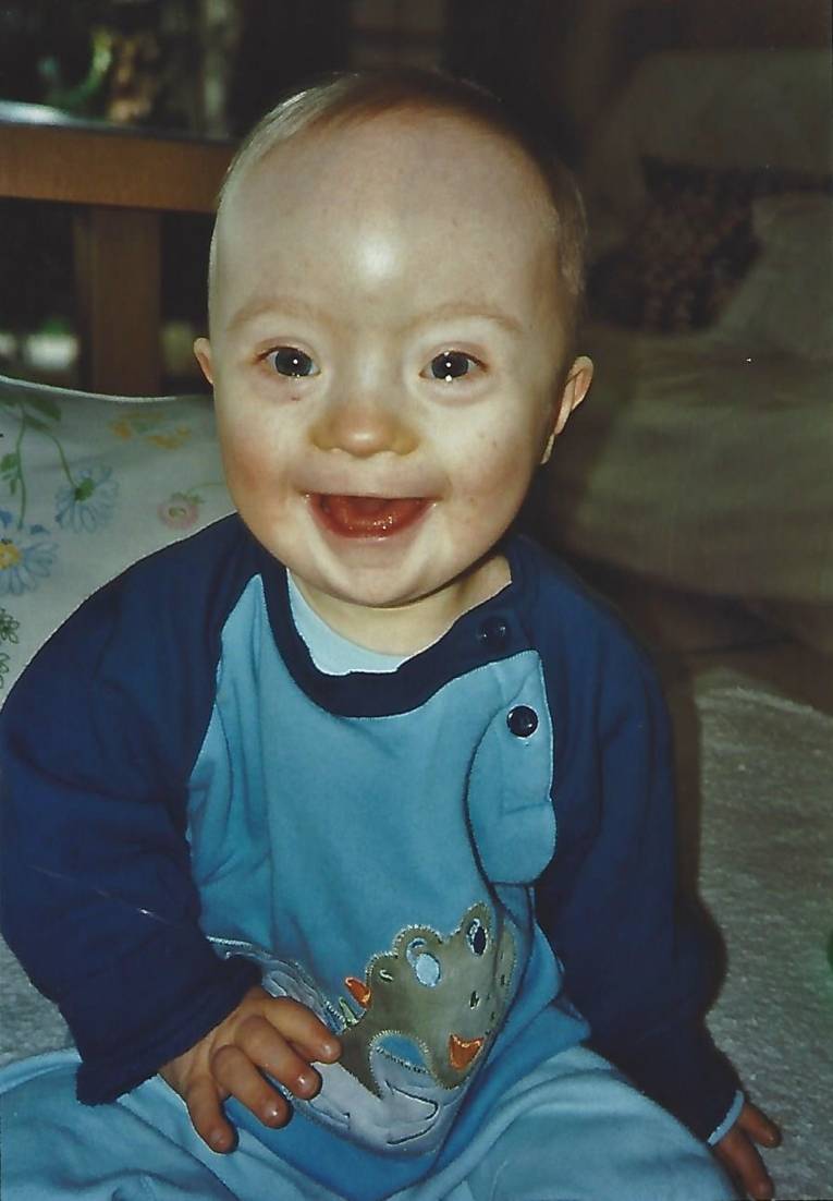 Farbfoto. Ein lachendes Baby im blauen Strampler sitzend auf ein Decke.